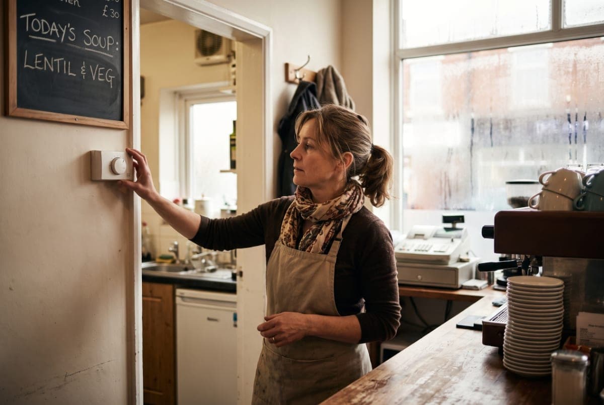 A café owner behind her counter at opening time, turning down the thermostat by the kitchen door.