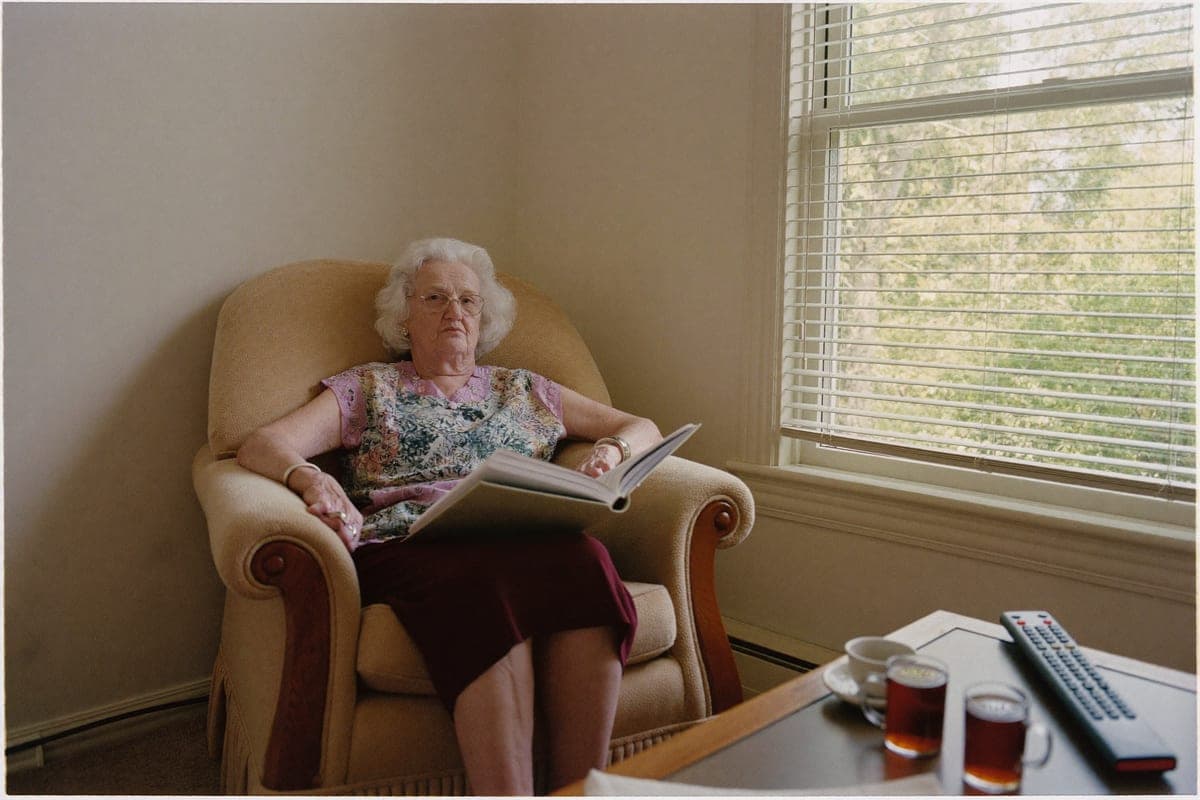 A woman in her seventies in her armchair by the window, a book open on her lap and a single-button remote on the table beside her tea.