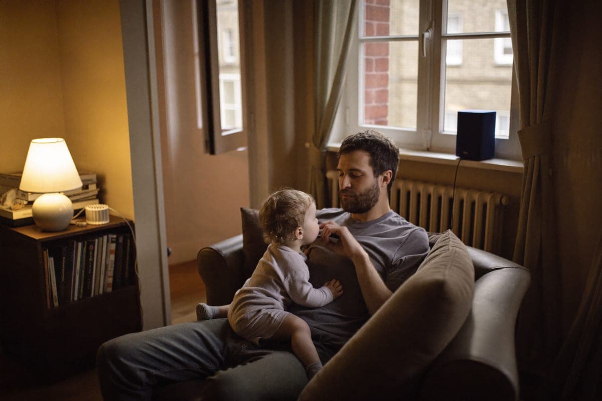 A father on the sofa with his toddler in a terraced-house living room in the evening, a table lamp on and a small speaker on the shelf behind them.